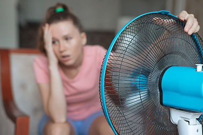 Young woman using electric fan at home in living room, sitting on couch cooling off during hot weather, suffering from heat, high temperature